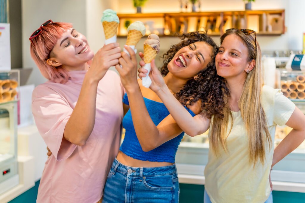 Drei junge Frauen stoßen in einer Eisdiele fröhlich mit ihren Eiswaffeln an.